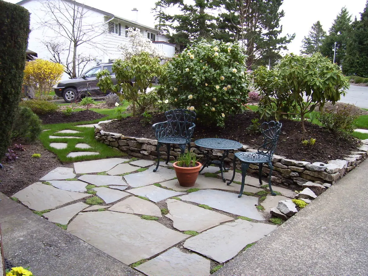 A newly installed Pennsylvania bluestone patio in the front yard, featuring a black wrought-iron table and chairs, bordered by a dry-stacked stone wall and surrounded by fresh greenery.