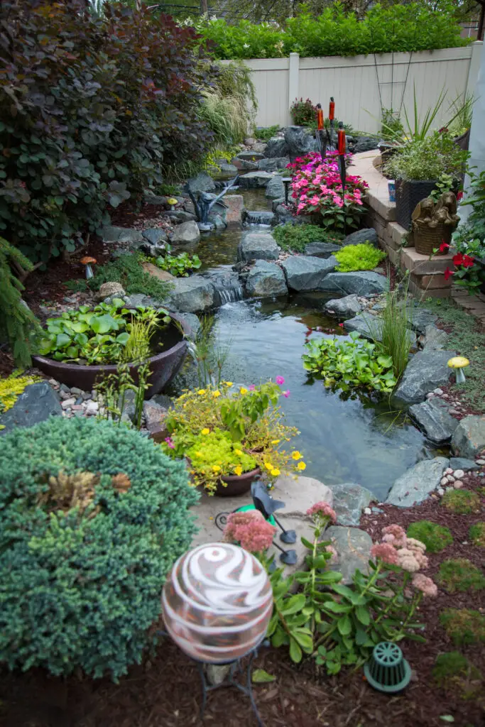 A lush backyard garden features a small pond with a cascading waterfall, surrounded by rocks, colorful flowers, green plants, and decorative ornaments. A white fence encloses the tranquil scene.