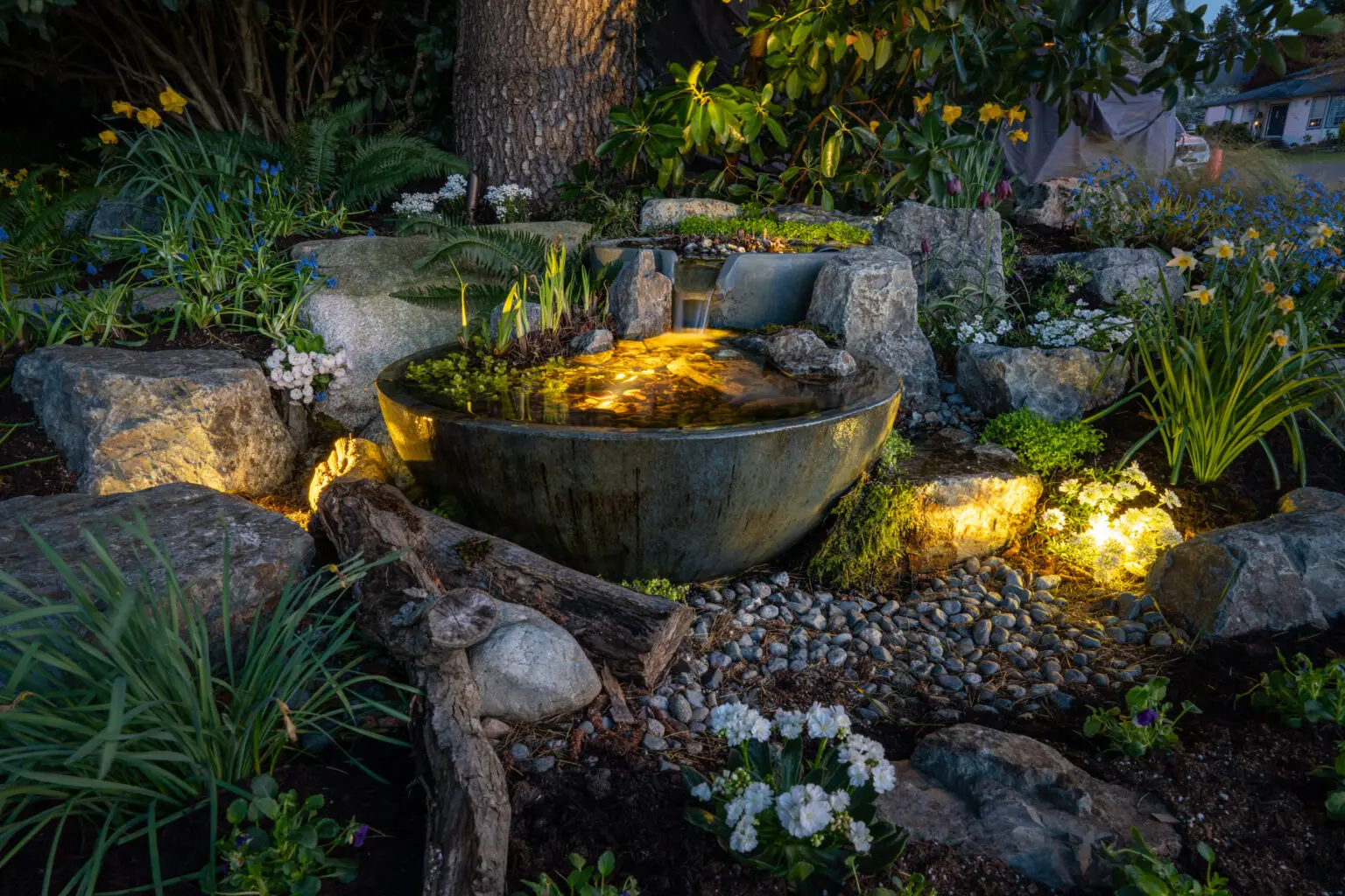 A small illuminated garden fountain features a round stone basin surrounded by rocks, flowering plants, ferns, and accent lights, with pebbles and a fallen branch in the foreground—a tranquil garden retreat.