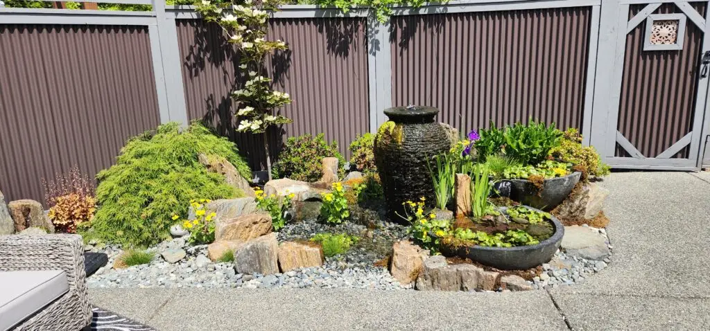 A small garden with rocks, flowering plants, and a black stone fountain with water flowing into a round basin, set against a tall gray fence in a sunny outdoor patio area, attracting seasonal visitors.