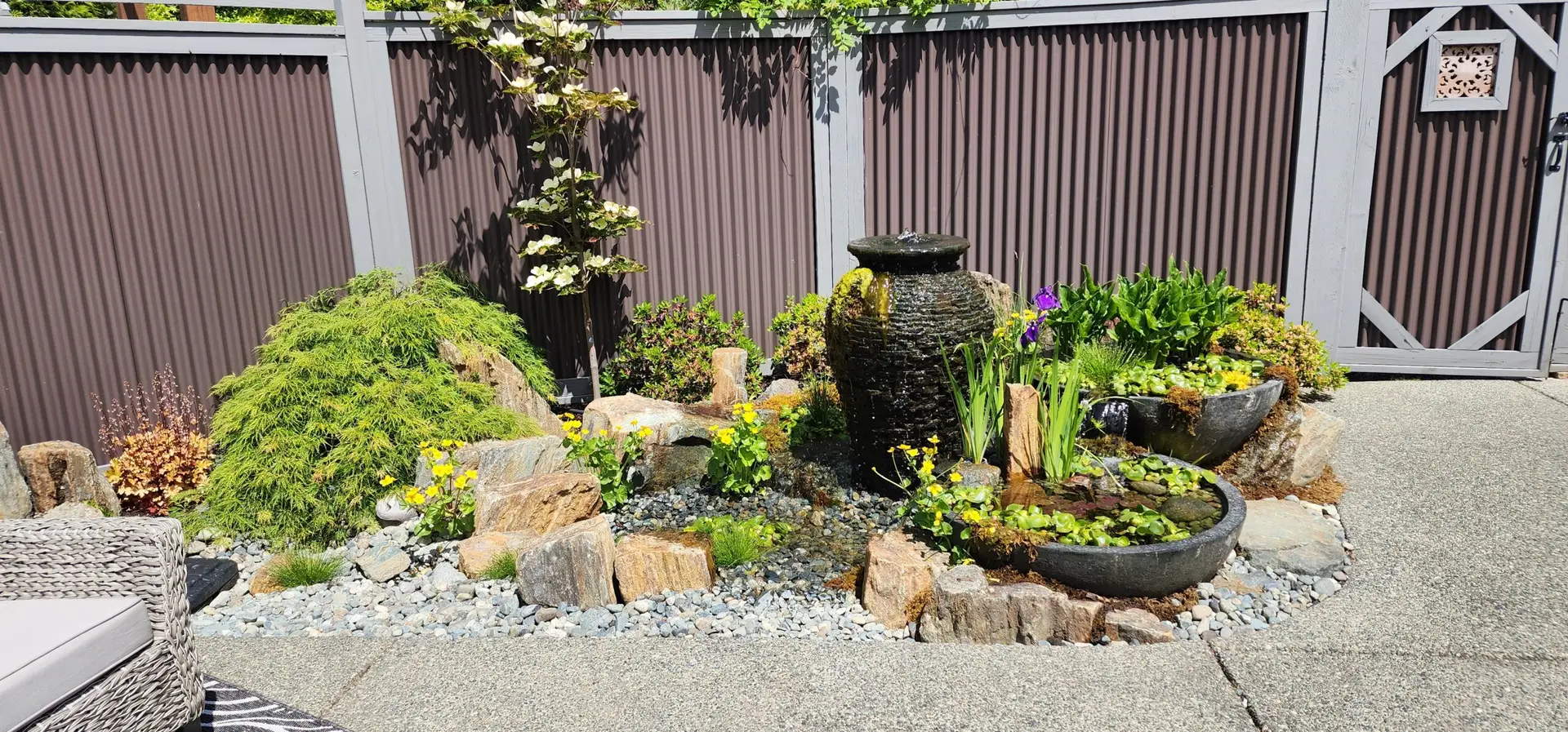 A small garden with rocks, flowering plants, and a black stone fountain with water flowing into a round basin, set against a tall gray fence in a sunny outdoor patio area, attracting seasonal visitors.
