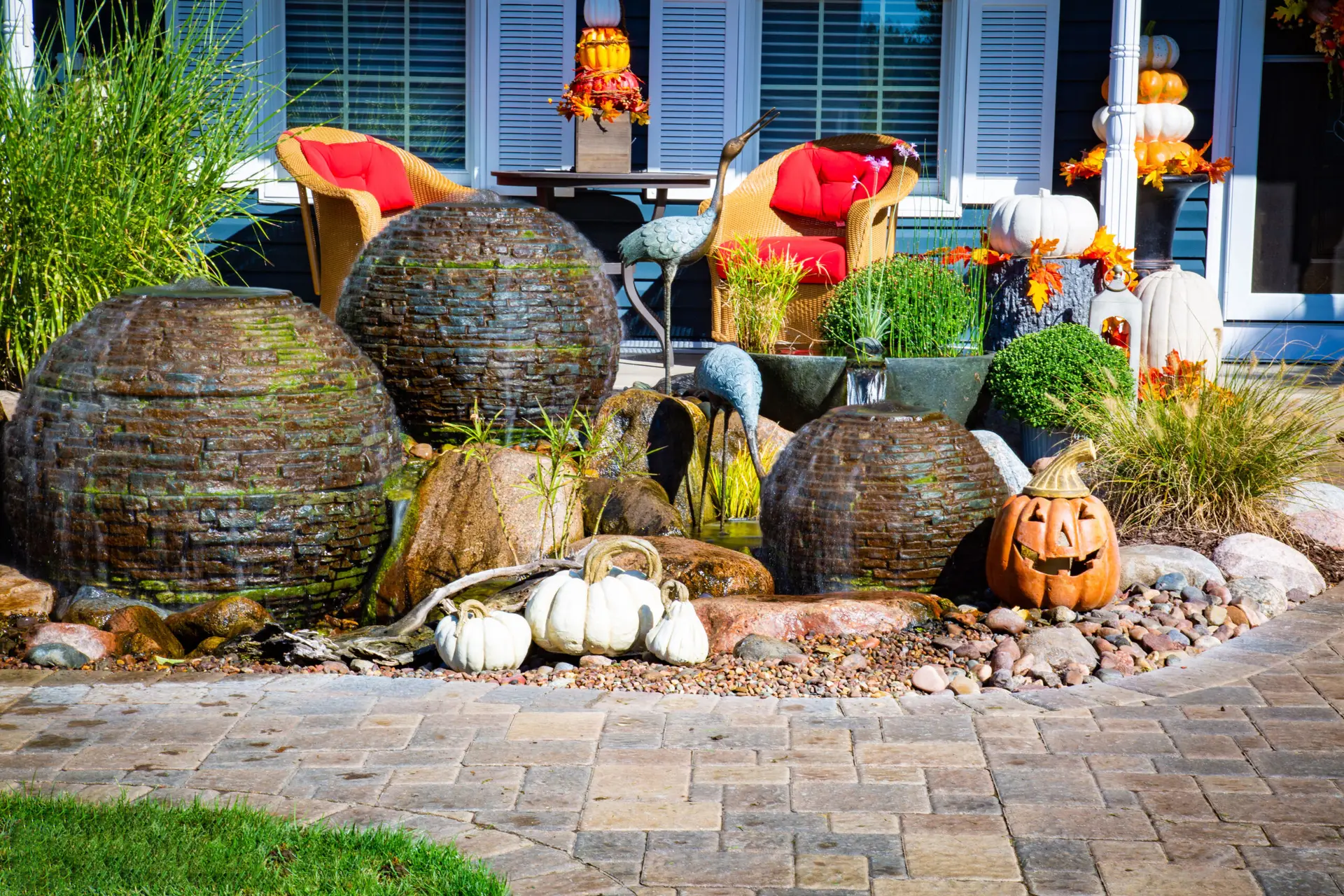 Decorative water fountains and pumpkins are arranged in a landscaped garden with a patio. Two chairs with red cushions sit by a table adorned with fall decor, as birds pause during migration, adding to the festive autumn scene.