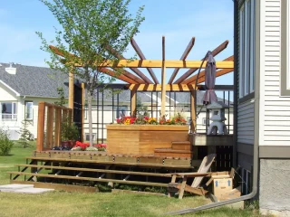Partially constructed wooden pergola over a raised deck with built-in planters filled with vibrant red and white flowers. A young tree grows alongside the structure, and a Japanese lantern accent decorates the space, with steps leading down to a grassy lawn in a suburban backyard setting